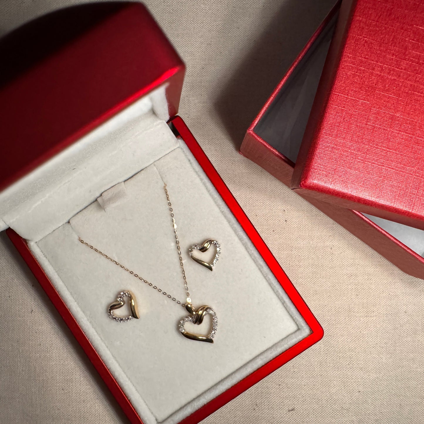 Image of the 10k Yellow gold heart set. The hearts have an intricate design with white diamond on the top right side of the heart and bottom left side of the heart. The heart is in a red and white box with the outer part of the box to the side.