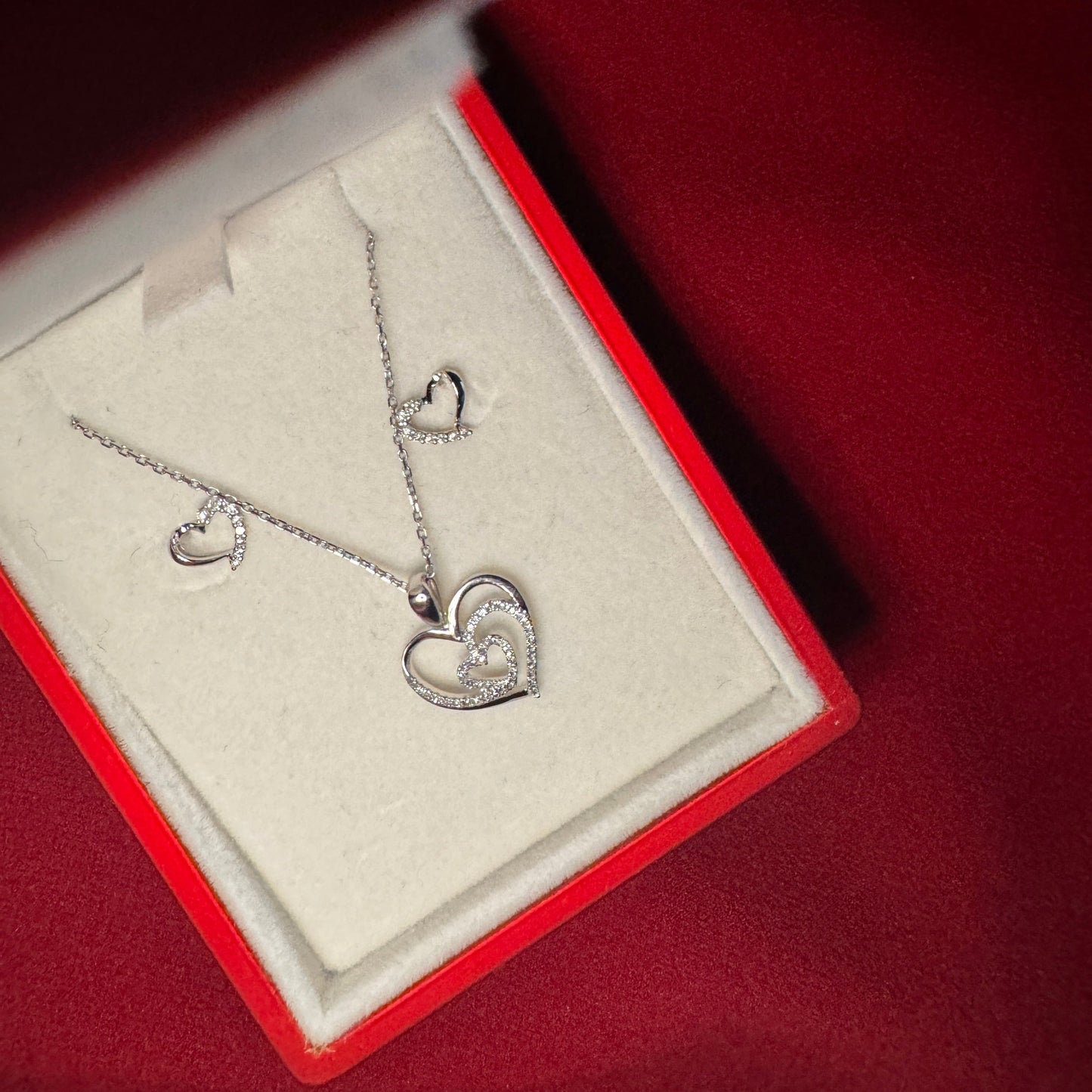 White gold heart set with white diamond. There are two heart studs with white accents on one side of the heart. And a heart pendant with a smaller diamond heart in the center. The set is in a red gift box with a red background.