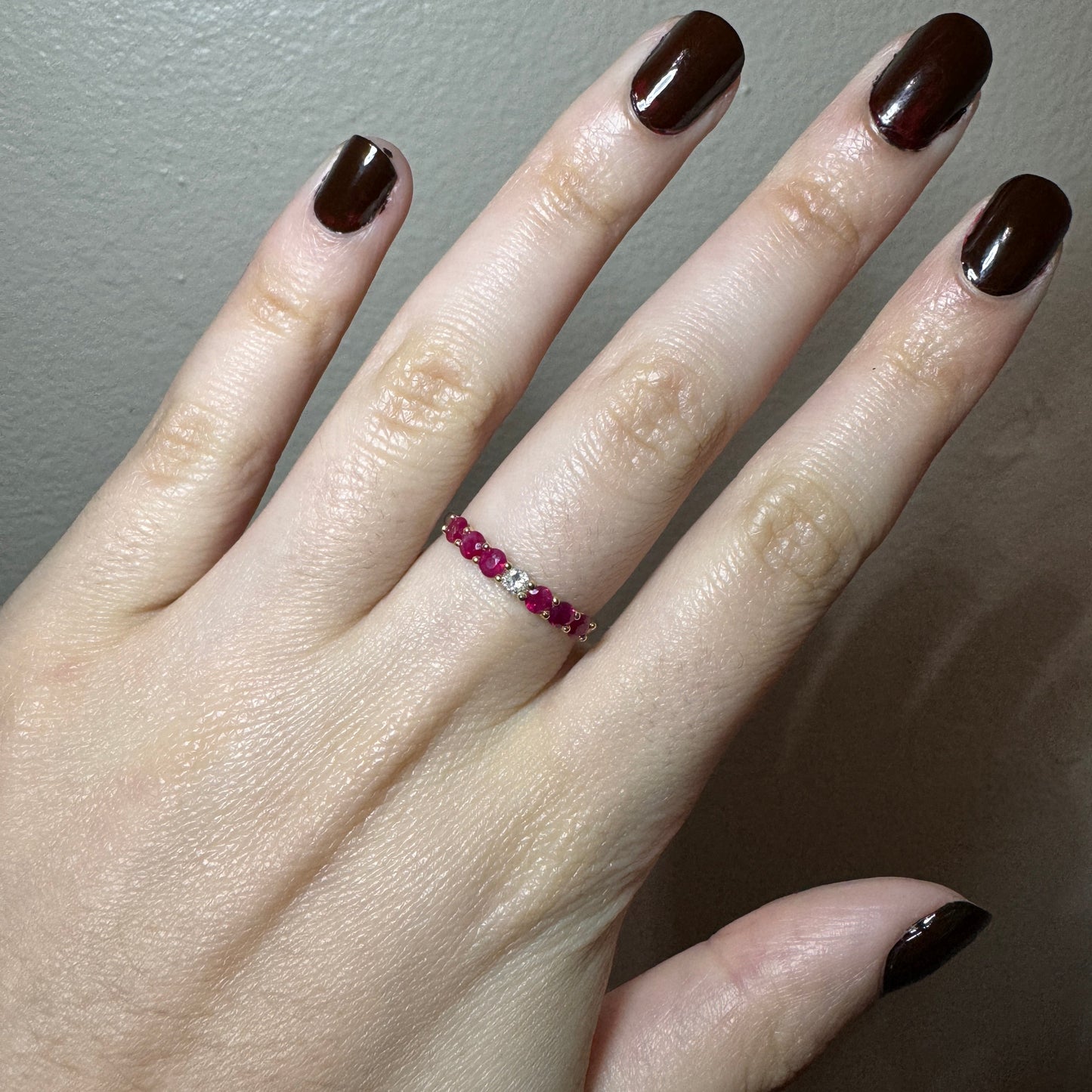 Model wearing white gold ring. The band is smooth. In the center of the ring there is one round white diamond. On each side of the diamond there are 3 round dark pink rubies. The rubies and the diamonds are held together by yellow gold prongs.