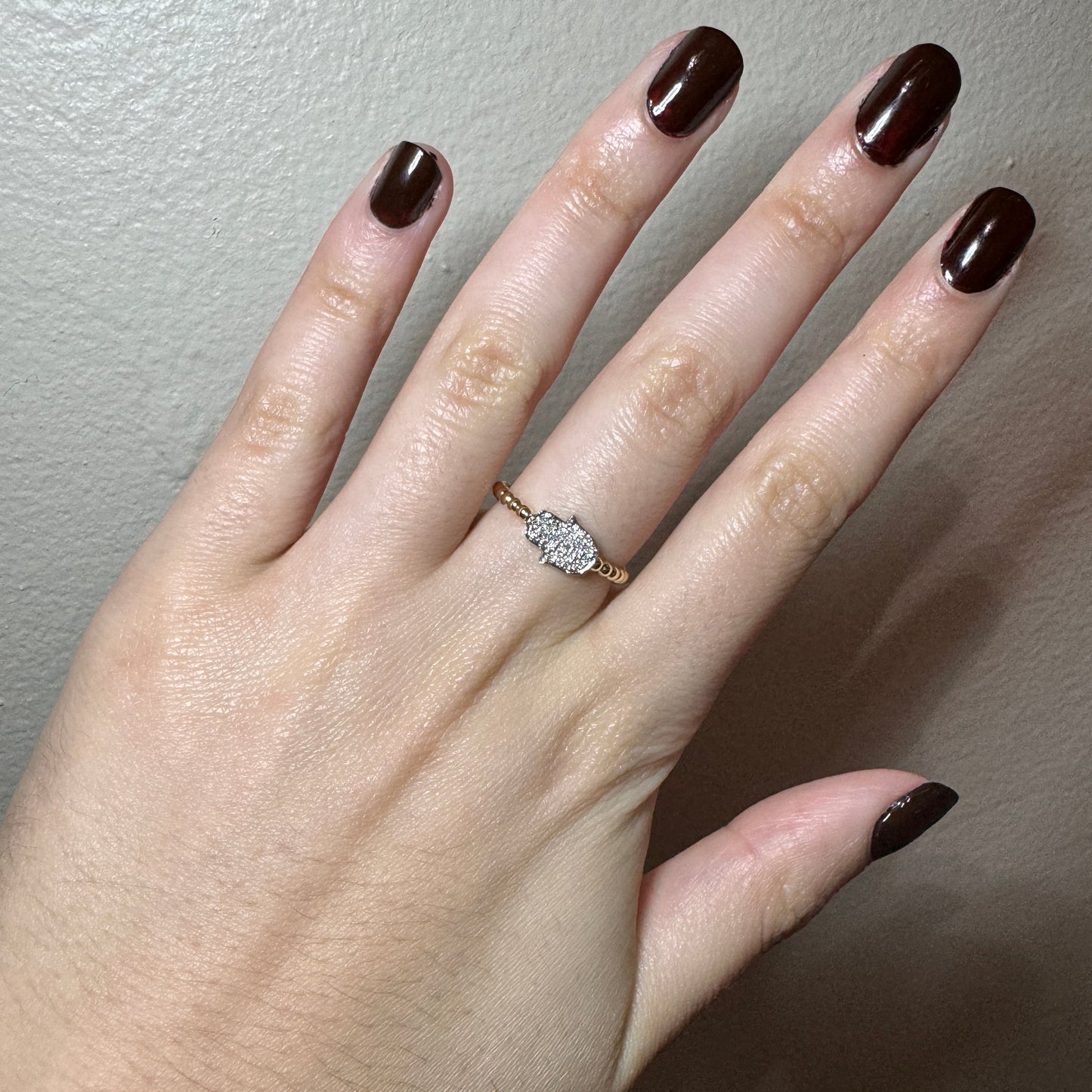 Model wearing yellow gold ring. The band of the ring has a circular design. In the center of the ring there is a horizontal white old hamsa hand. The hand is filled with small round white diamonds.