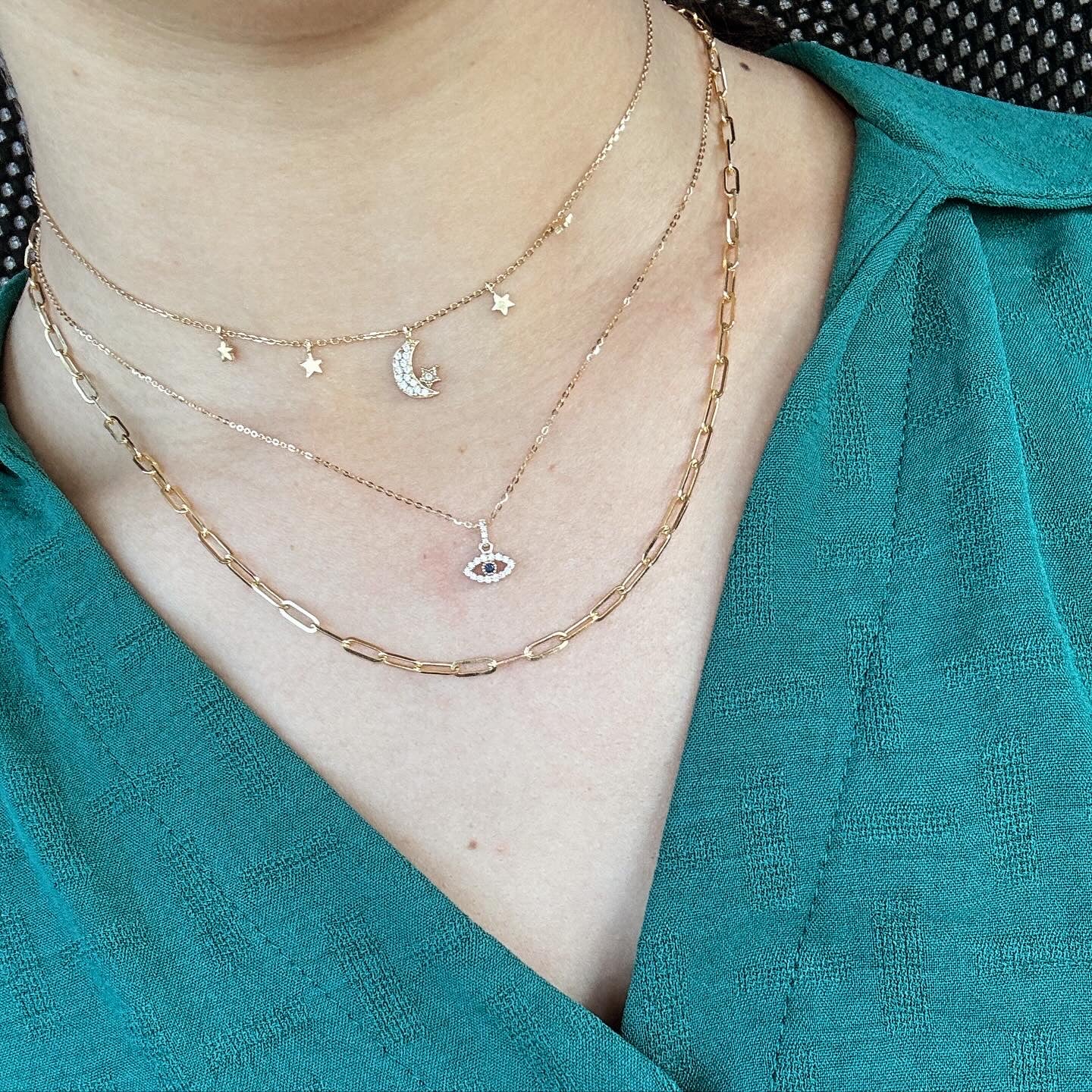 Model wearing 3 yellow gold necklaces. The first one has a crescent moon with two stars on either side. The second necklace is the yellow gold eye pendant outlined with white diamonds and a blue sapphire in the center. The last necklaces is a plain paperclip chain.