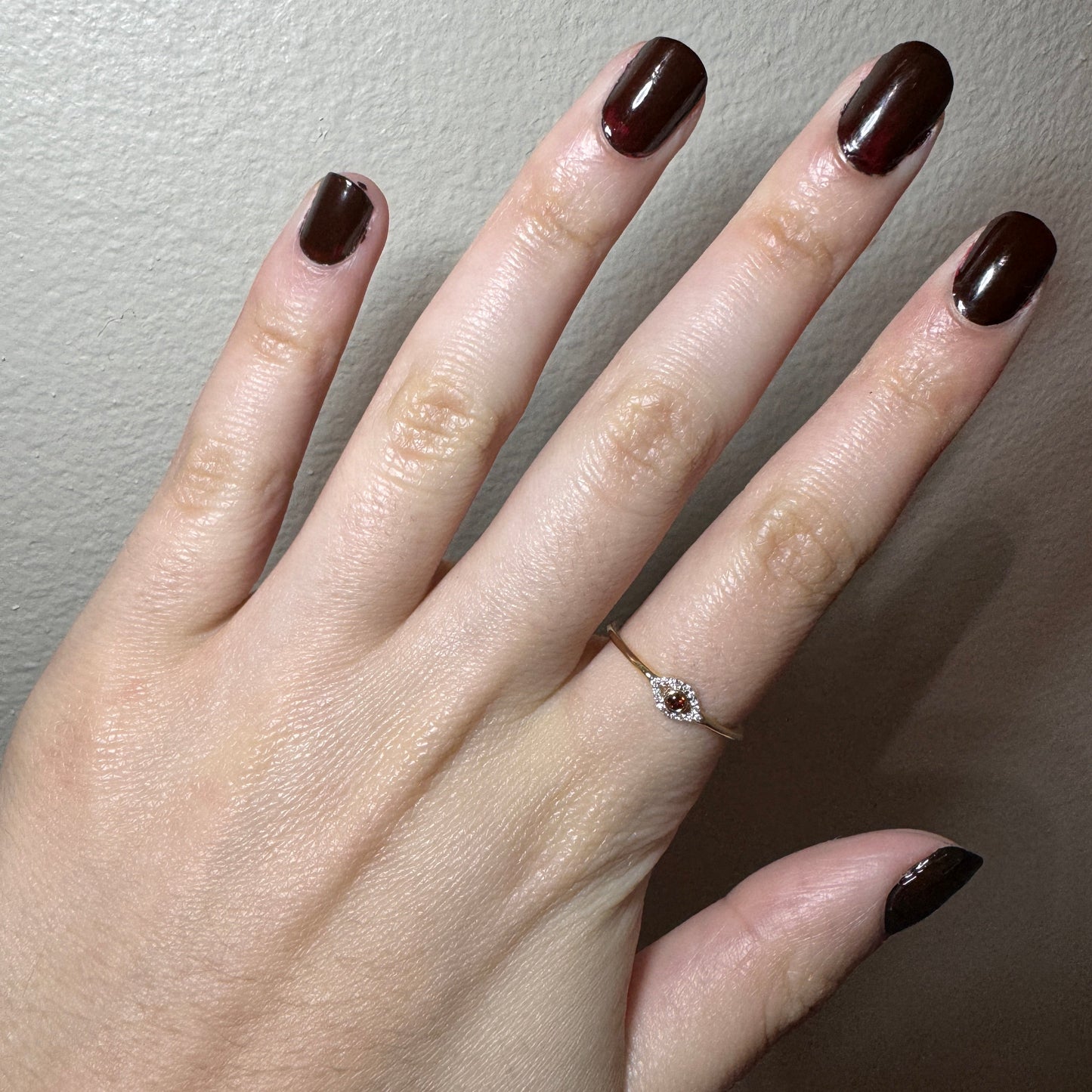 Model wearing yellow gold ring. The ring has a smooth band. In the center there is an eye shape. The outline of the eye has small white diamonds. The center of the eye has a dark almost wine color ruby with a thin yellow gold outline.