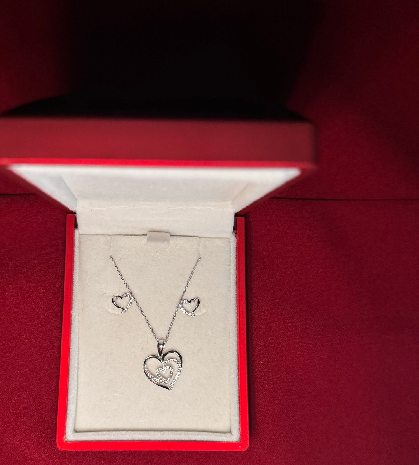 White gold heart set with white diamond. There are two heart studs with white accents on one side of the heart. And a heart pendant with a smaller diamond heart in the center. The set is in a red gift box with a red background.