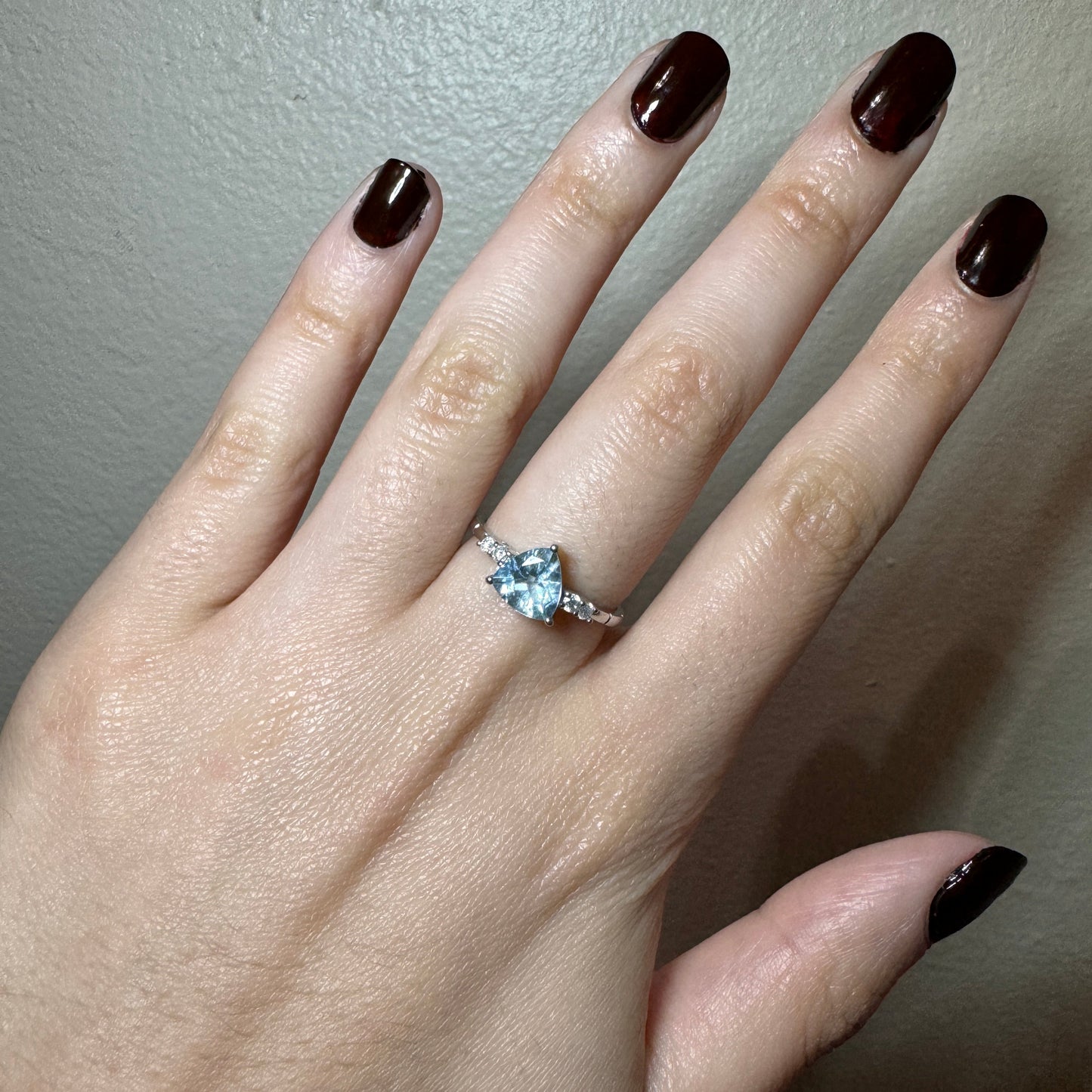 Model wearing white gold ring. The ring has a triangular light blue aquamarine stone in the center. On each side of the stone there are2 small round white diamonds.