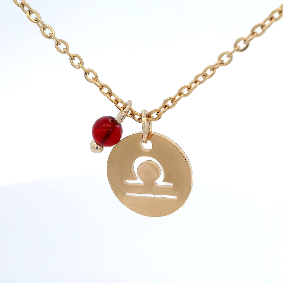 A gold-plated stainless steel chain necklace with a round gold disc pendant featuring a cutout symbol of the Libra zodiac sign, adorned with a small bead, on a white background.