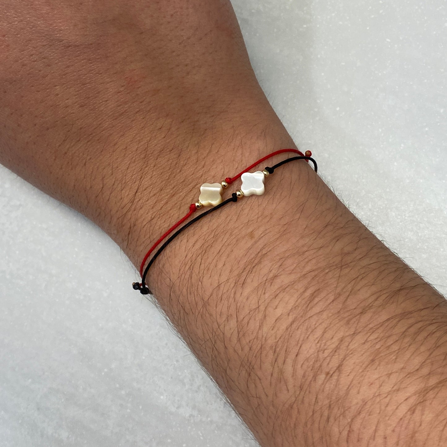 Model wearing two handmade adjustable bracelets with a Mother of Pearl clover in the center and 2 gold beads on each side. One bracelet has s a red string and the other has black string