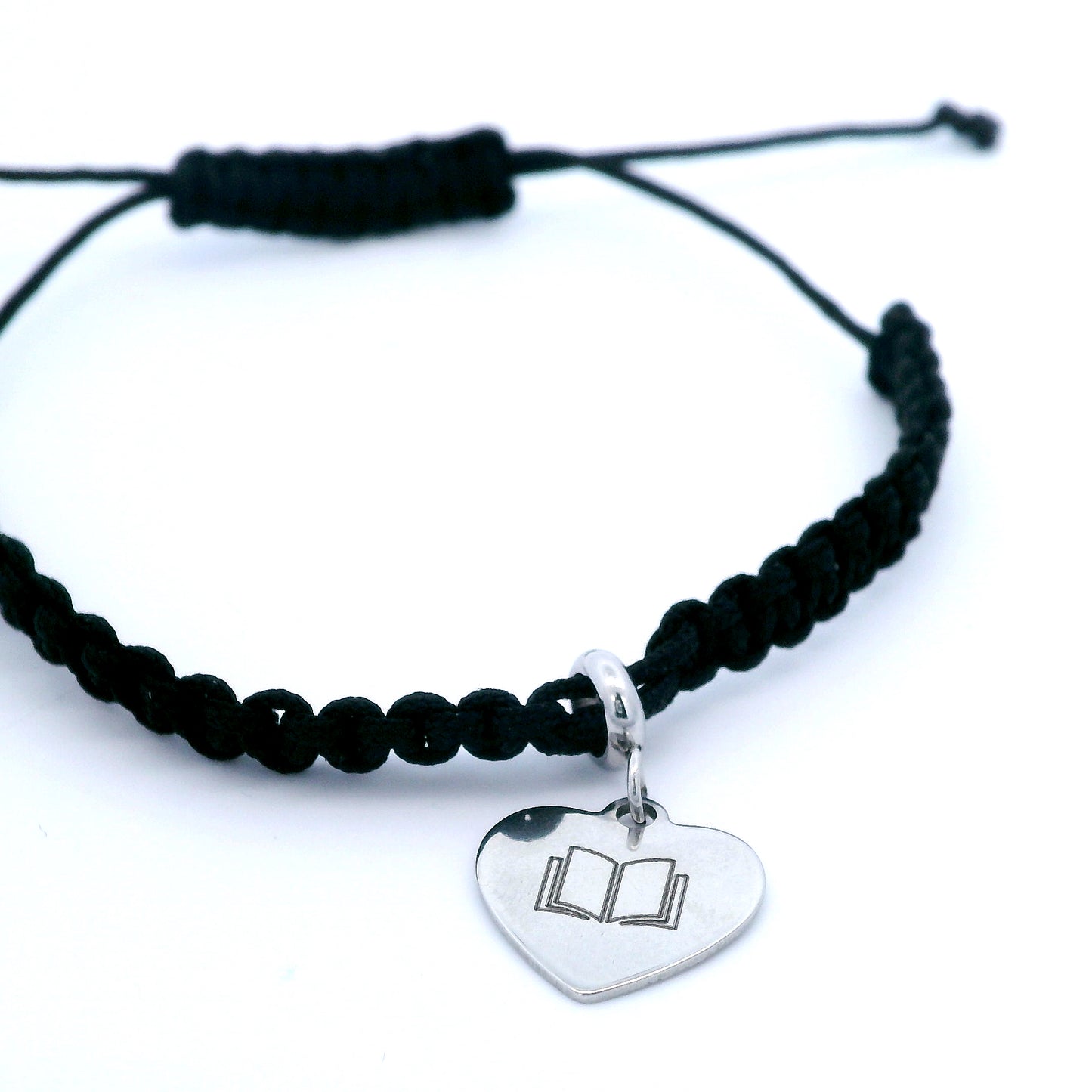 A black macrame bracelet bracelet with a silver heart-shaped charm engraved with an open book illustration, shown on a clean white background.