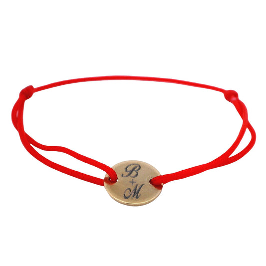 Red string bracelet with a round yellow stainless steel featuring the initials "B" and "M" with a plus sign in the middle on a white background