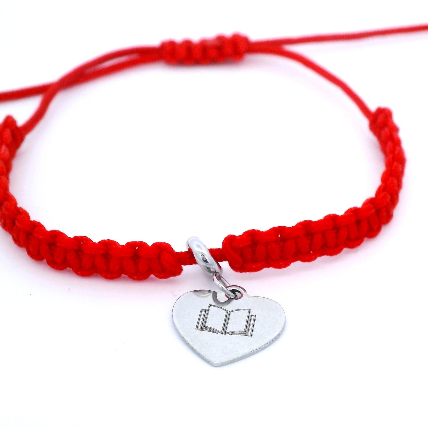 A red macrame bracelet with a silver heart-shaped charm engraved with an open book illustration shown on a clean white background.