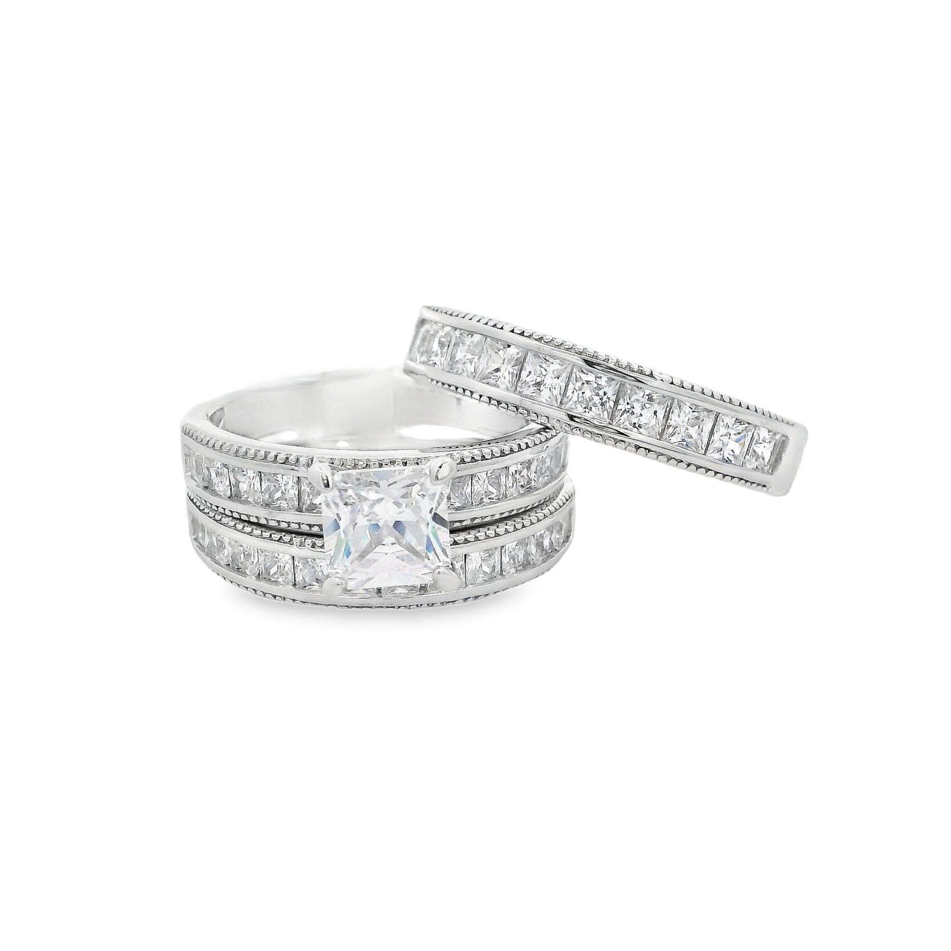 Front view of Silver bridal trio set. On the bottom there is a wedding band with square zirconia. On top is an engagement ring with a square zirconia in the center and smaller square zirconia on the sides. Laying on top at an angle is another wedding band that is thicker and with square zirconia.