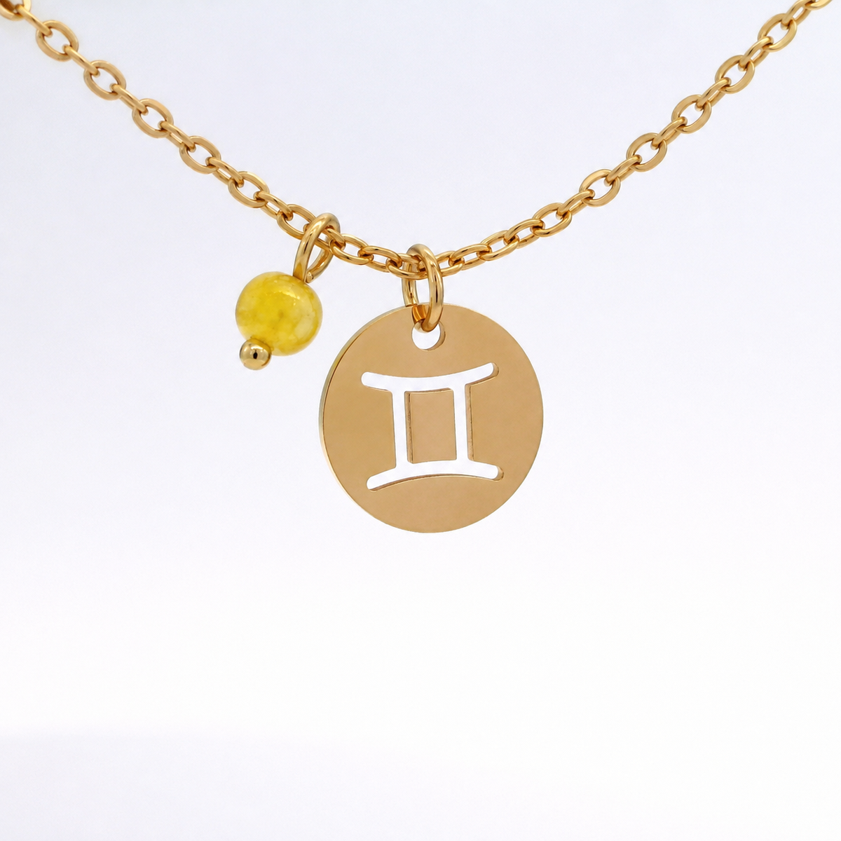 A gold-plated stainless steel chain necklace with a round gold disc pendant featuring a cutout symbol of the Gemini zodiac sign, adorned with a small bead, on a white background.